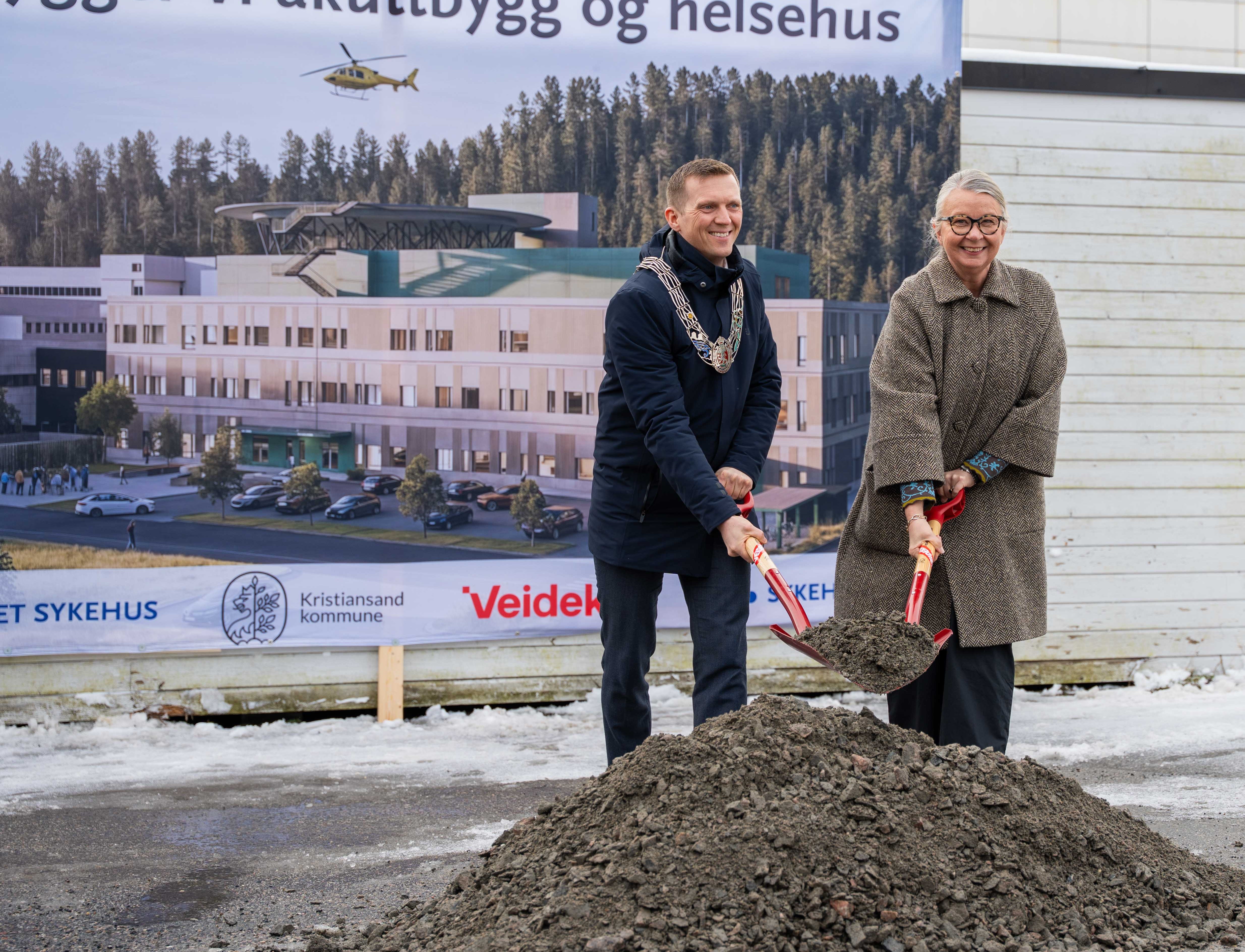 En mann og en kvinne som st&aring;r ved en jordhaug med spade foran et stort banner med bilde av nytt akuttbygg