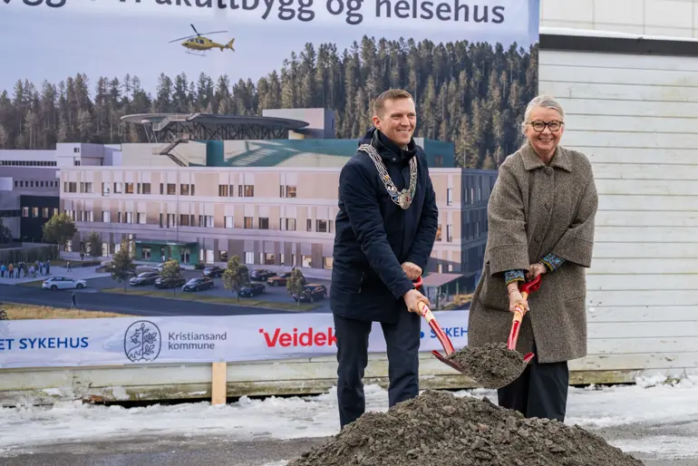 En mann og en kvinne som står ved en jordhaug med spade foran et stort banner med bilde av nytt akuttbygg