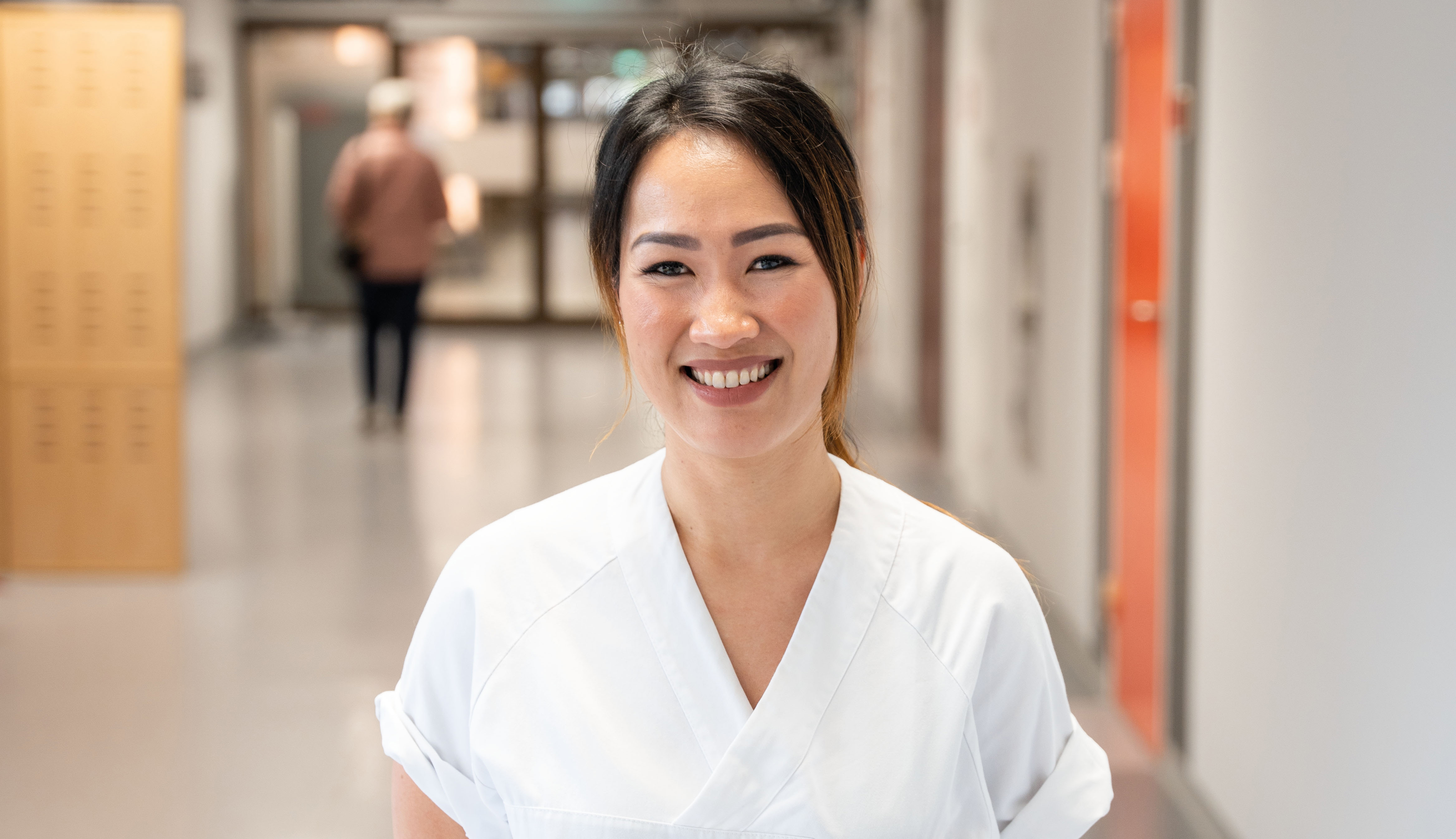 Sykepleier kledd i hvit uniform st&aring;r smilende i en korridor p&aring; sykehuset.