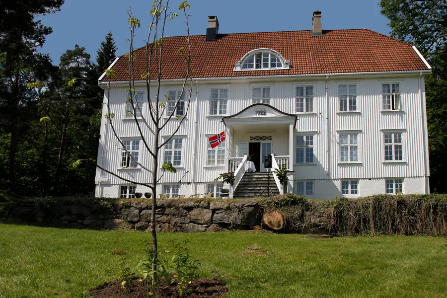 Hvitt trebygg liggende i grønne omgivelser på Eg. Foto.